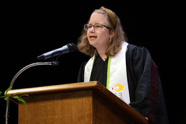 Ms. Mary White speaks from behind a podium at the Helen Keller School of Alabama Graduation. She wears her black ceremonial robes, a green cord, and a white stole. 